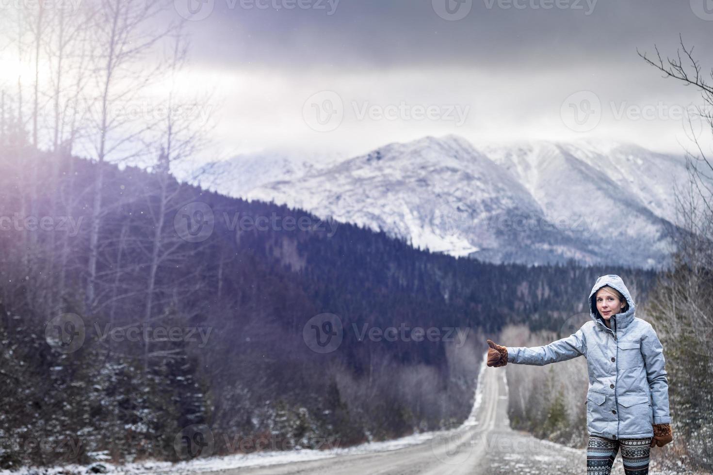 Mujer haciendo autostop en una carretera de invierno con hermosas montañas nevadas en segundo plano. foto