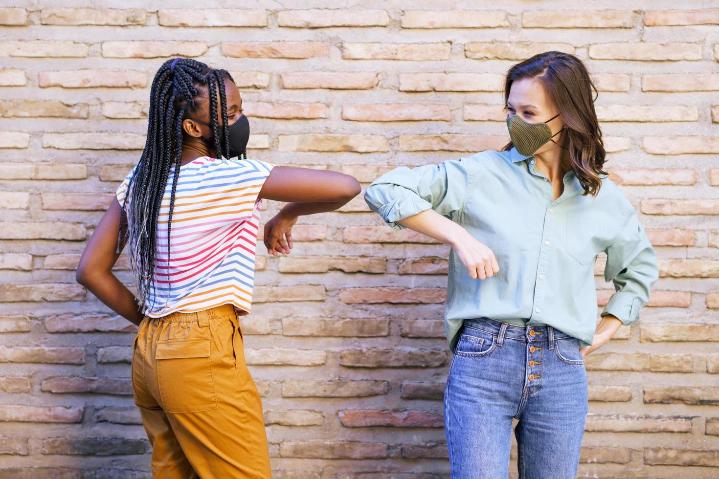 Multiethnic young women wearing masks greeting at each other with their elbows photo