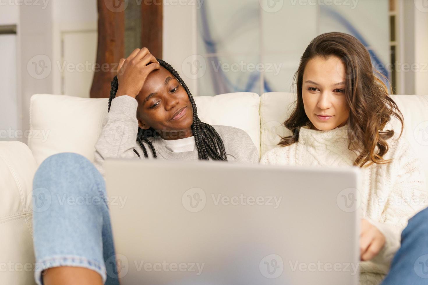 Two female student friends sitting on the couch at home using a laptop. photo