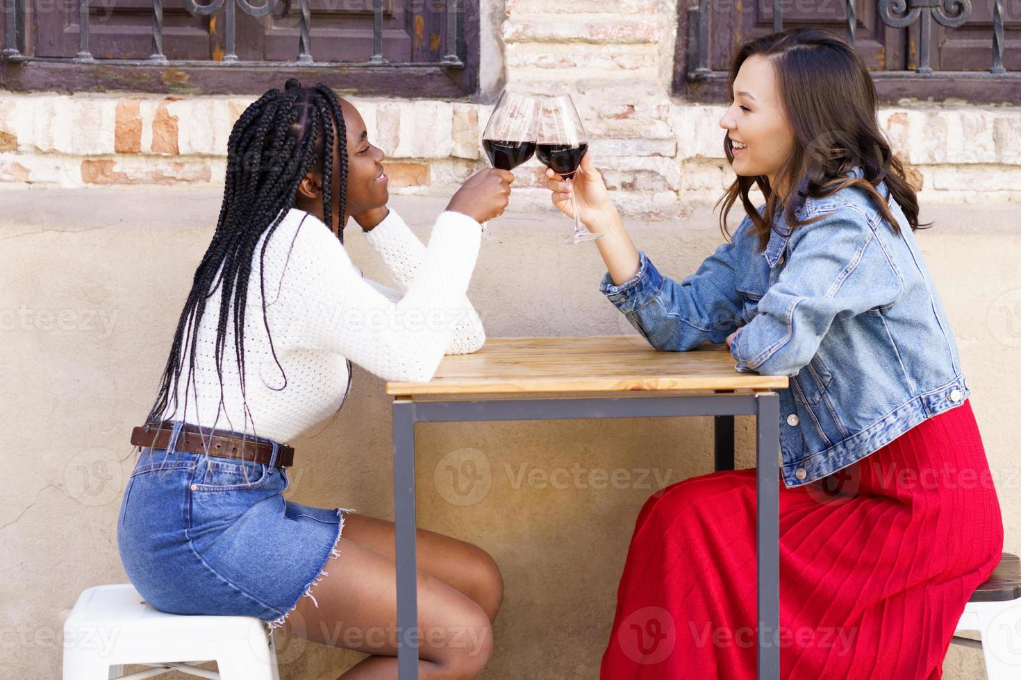 Two women making a toast with red wine sitting at a table outside a bar. photo