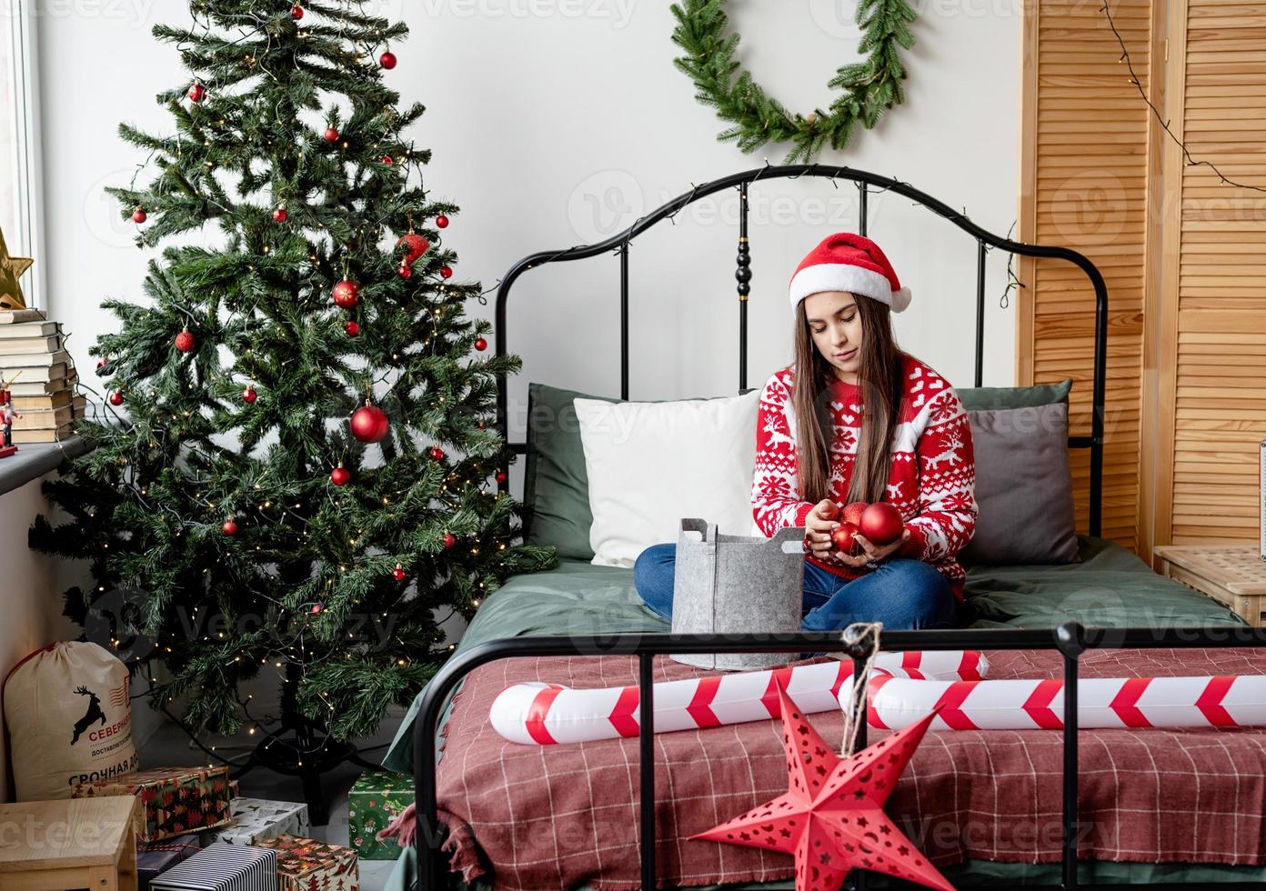 Mujer joven en suéter rojo y gorro de Papá Noel sentado en la cama celebrando la Navidad foto