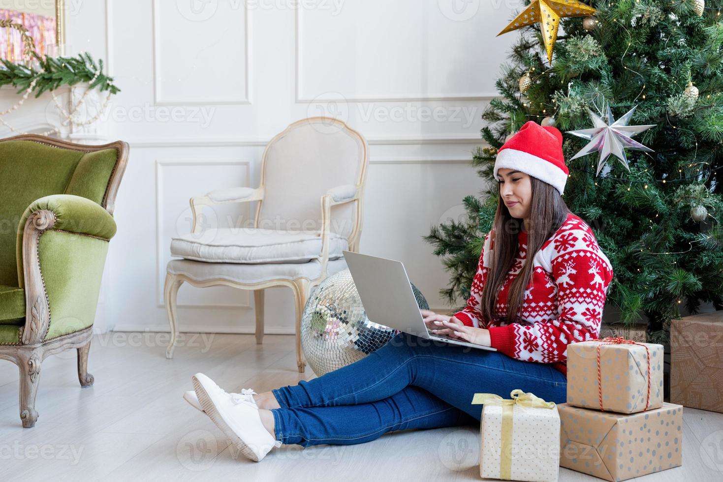 Mujer joven sentada cerca del árbol de Navidad en una sala de estar decorada trabajando en una computadora portátil foto