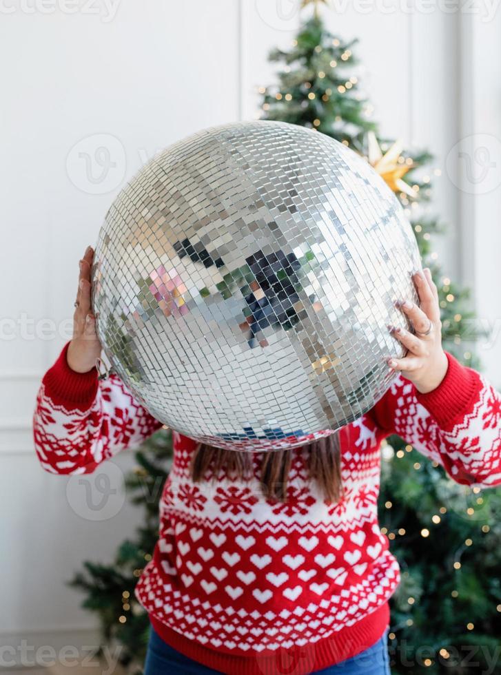 Young woman in santa hat holding mirror disco ball standing by the Christmas tree photo