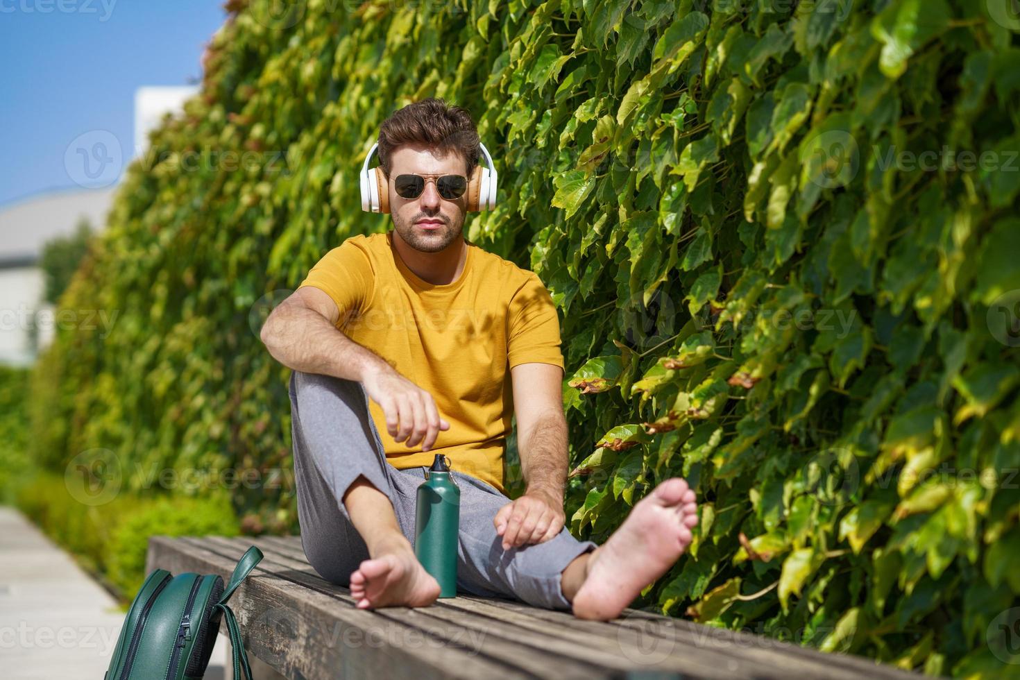 Male sitting outside using an aluminum water bottle, headphones and backpack. photo