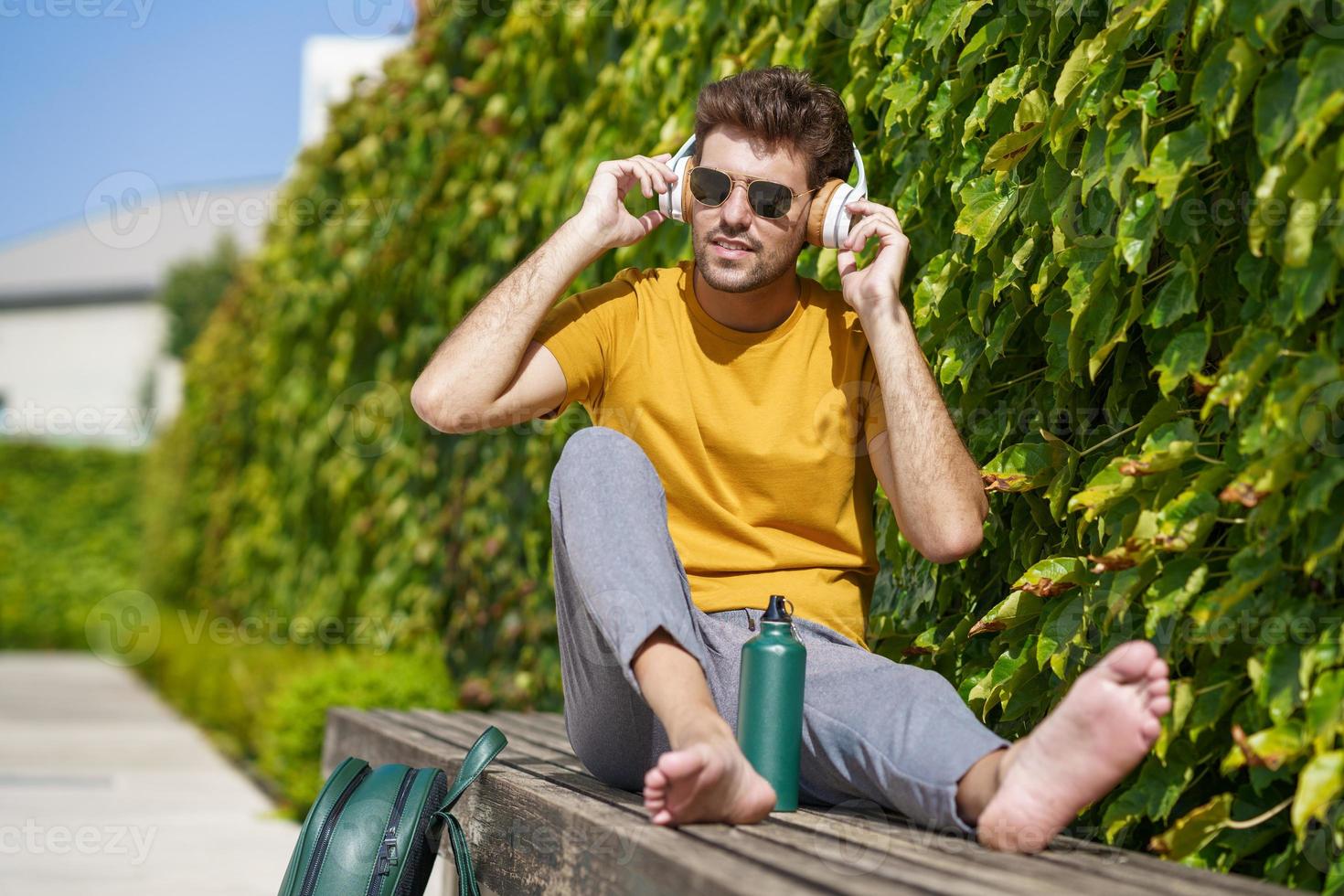 Male sitting outside using an aluminum water bottle, headphones and backpack. photo