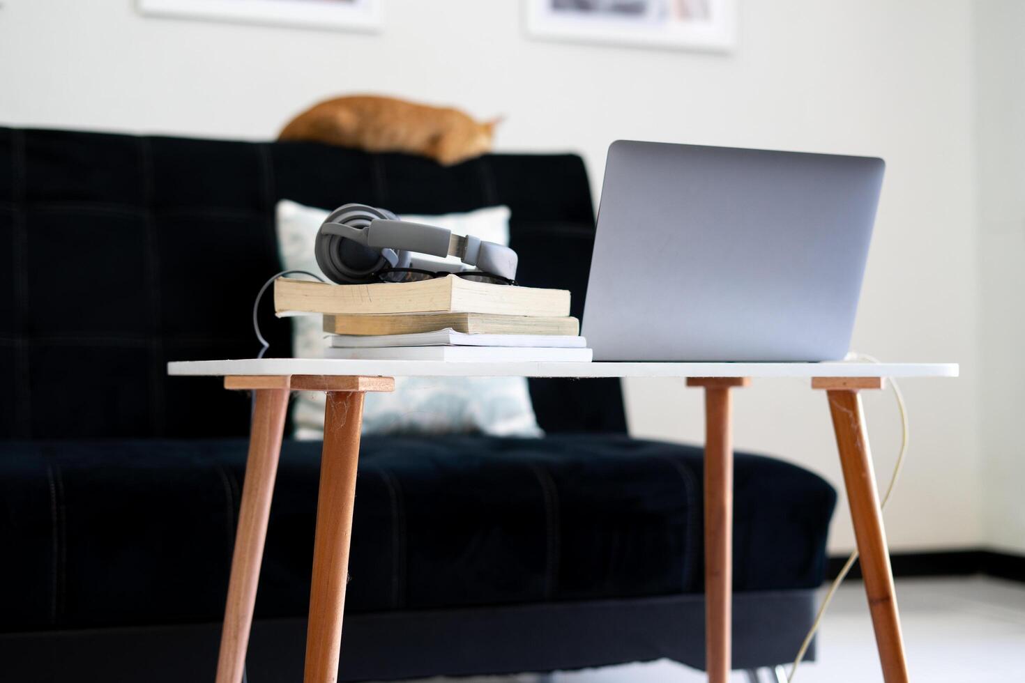 a laptop, a pile of books, and earphones on a white table. and the orange cat sleeping on the black sofa. photo