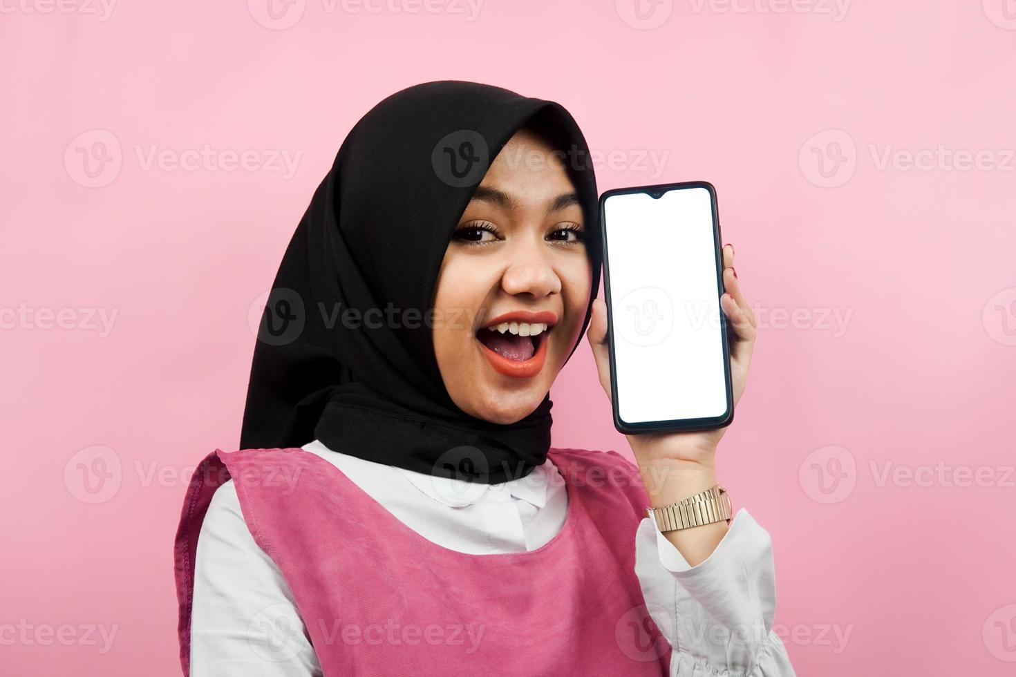 Close-up of beautiful and cheerful young Muslim woman holding smartphone with white or blank screen, promoting app, promoting something, isolated, advertising concept photo