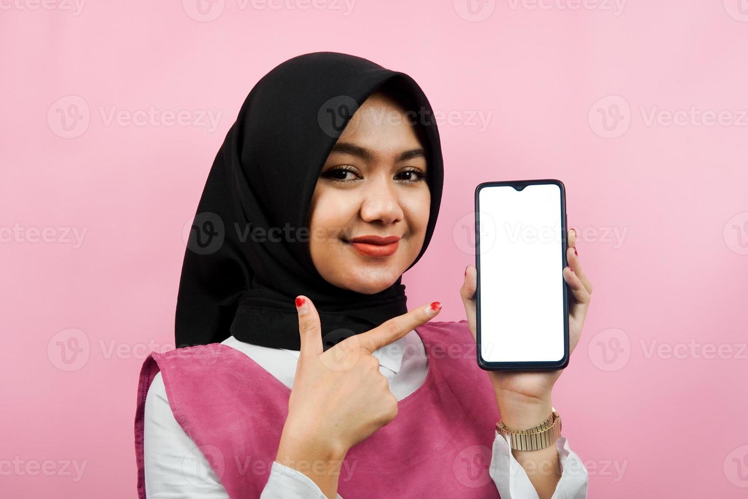 Close-up of beautiful and cheerful young Muslim woman holding smartphone with white or blank screen, promoting app, promoting something, isolated, advertising concept photo