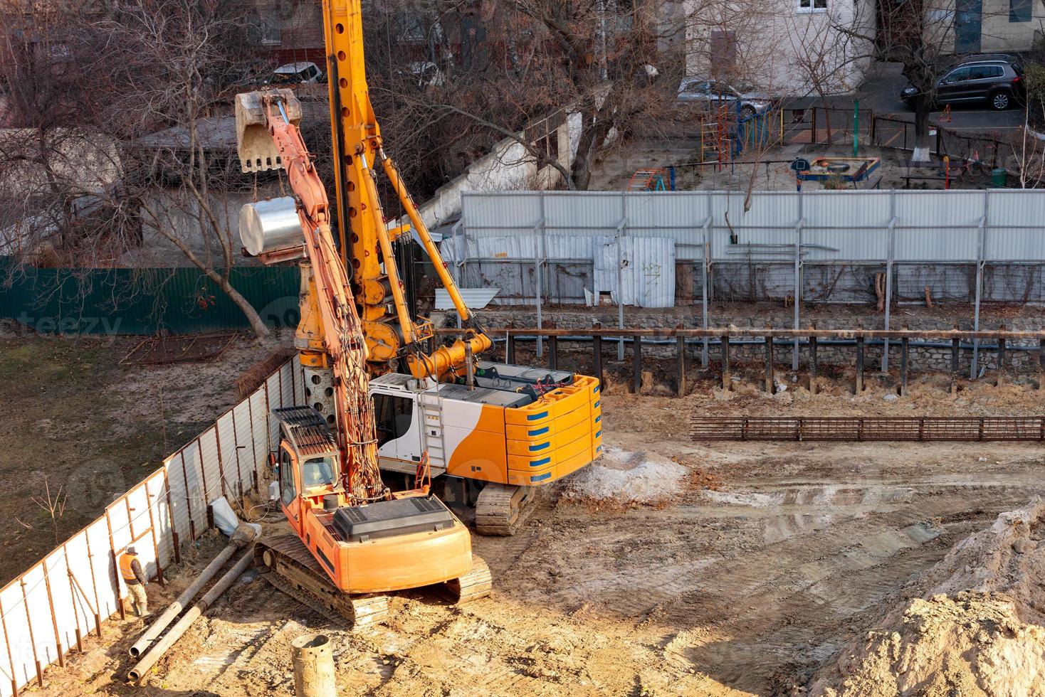 El equipo de construcción pesado montado sobre orugas trabaja en un sitio de construcción cercado entre edificios residenciales en una calle de la ciudad. foto