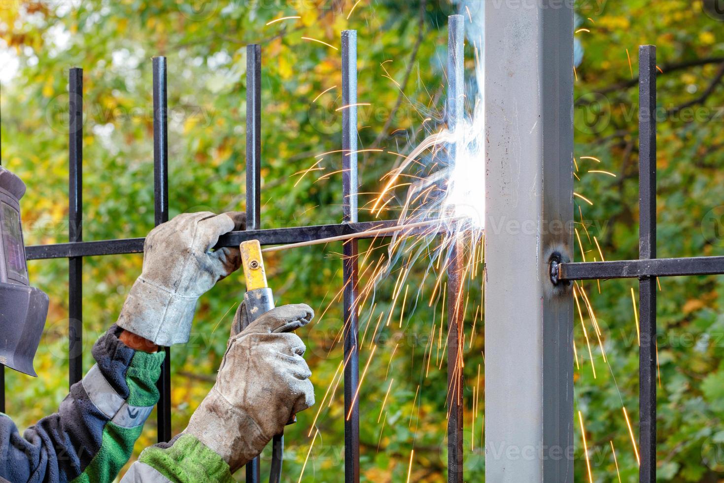 A welder in a safety helmet and gloves welds an iron grate to a metal post against a blurred background of a green park. photo