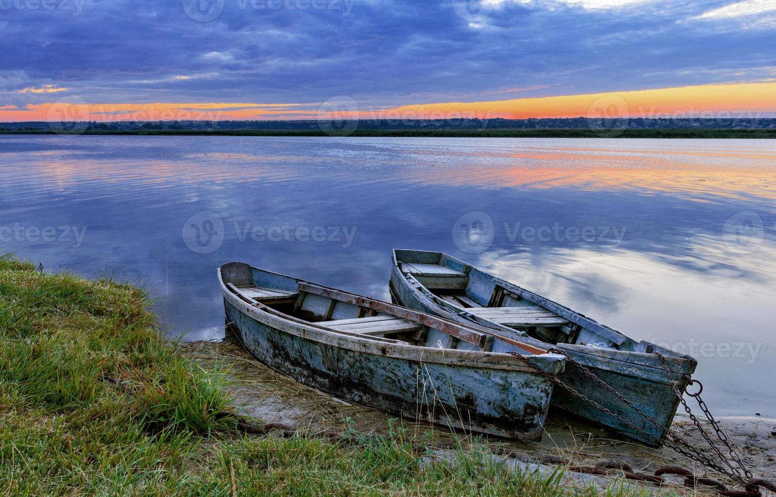 Two old dilapidated boats are moored with a metal chain to the bank of a calm river in the early morning against the backdrop of a cloudy sky and the rising sun. photo