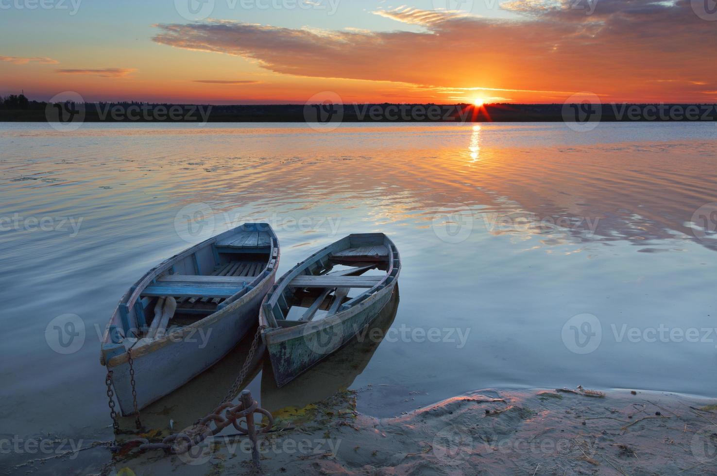 Two old blue-green boats stand on the bank of a calm river against the background of a bright rising sun photo