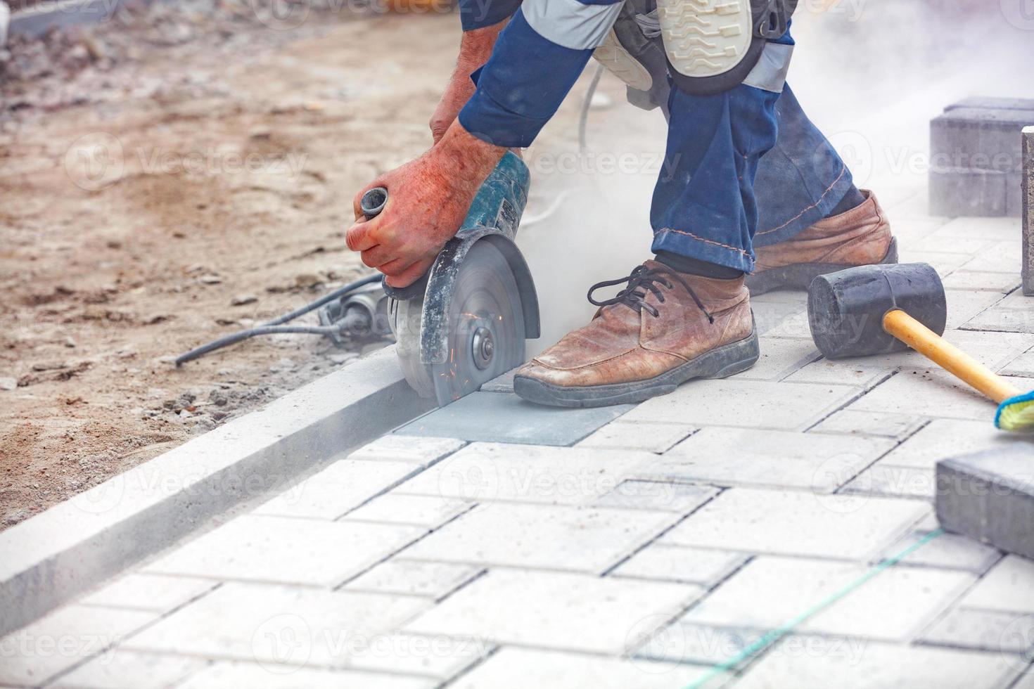 The hands of a builder with the help of a grinder cut the paving slabs against the backdrop of the work site. Close-up, blurred background. photo