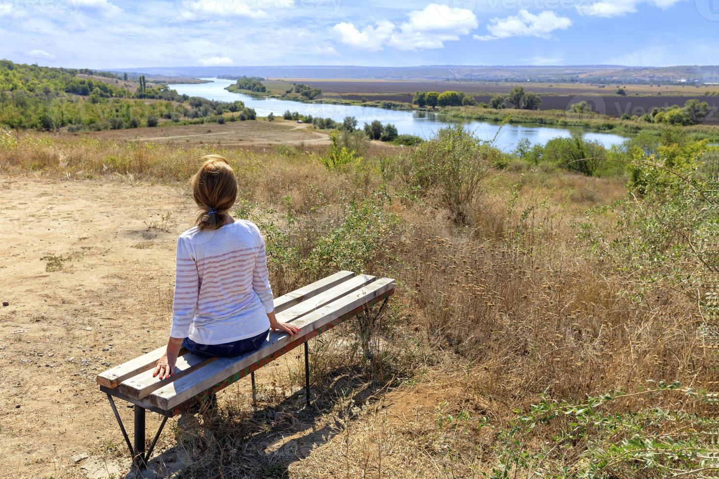 A young woman sits on a wooden bench and looks at the calm Southern Bug River on a sunny autumn day. photo