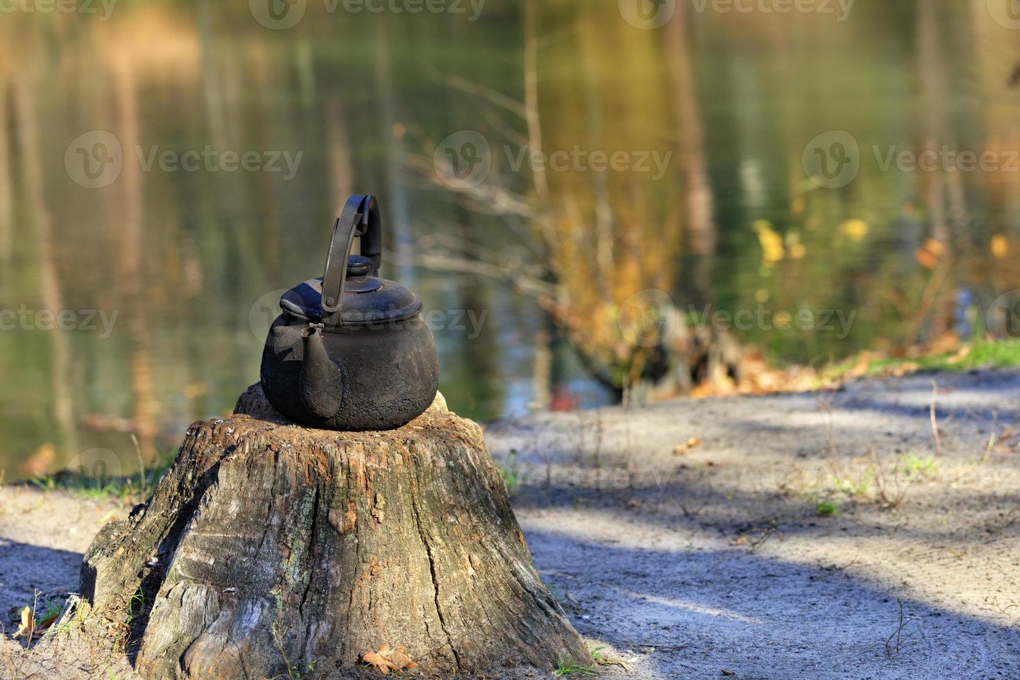 Metal burnt kettle stands on an old stump in the open air against a background of a forest lake. photo