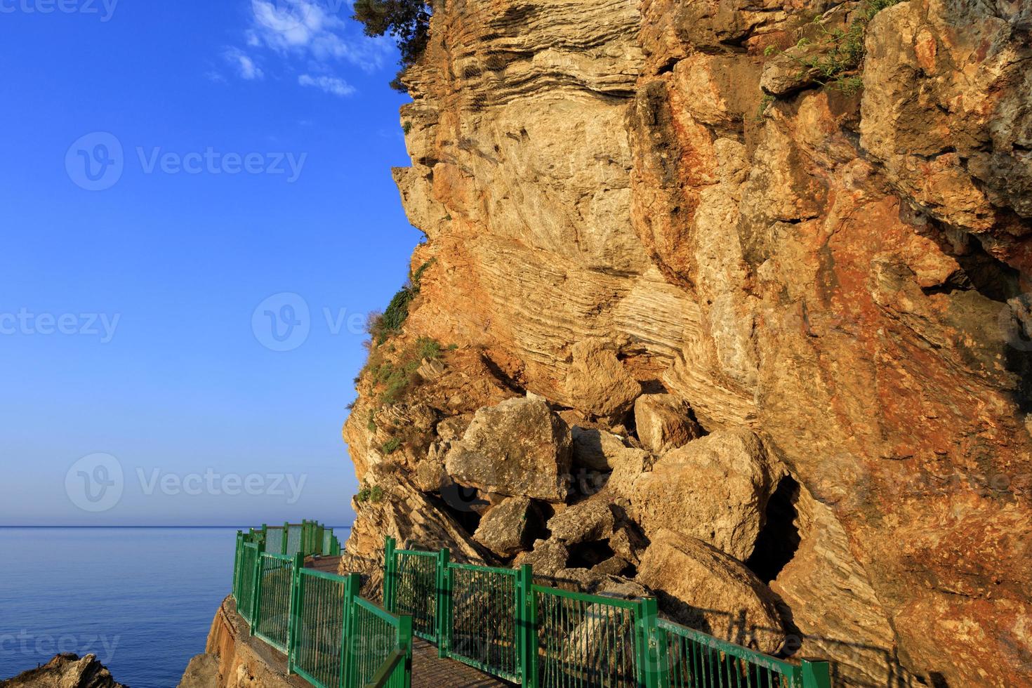 The road along the sea coast, surrounded by a green metal fence, is illuminated by the rays of the morning golden sun near a rocky cliff. photo