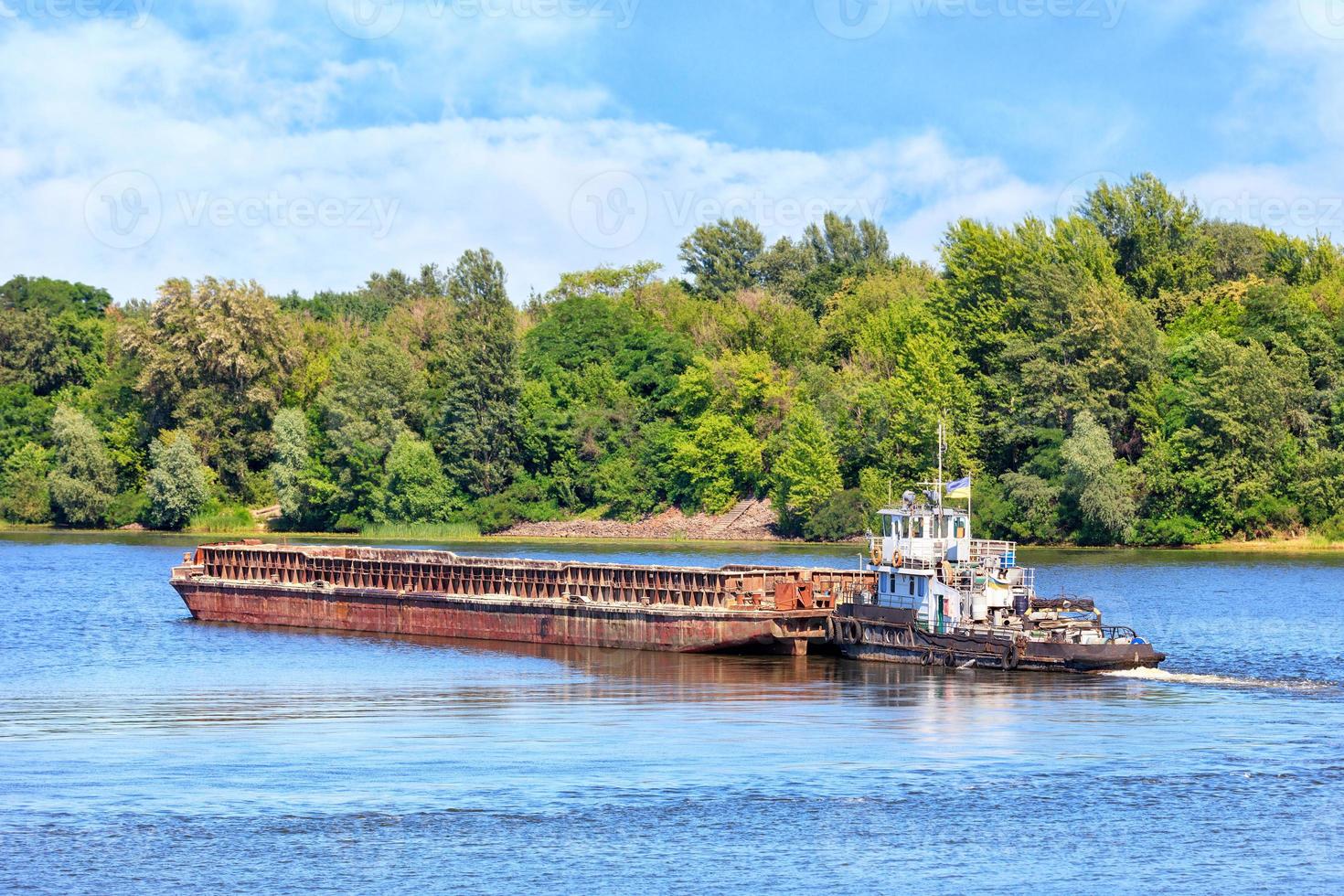 A river tug is pushing a rusty barge along the river along the shore. photo
