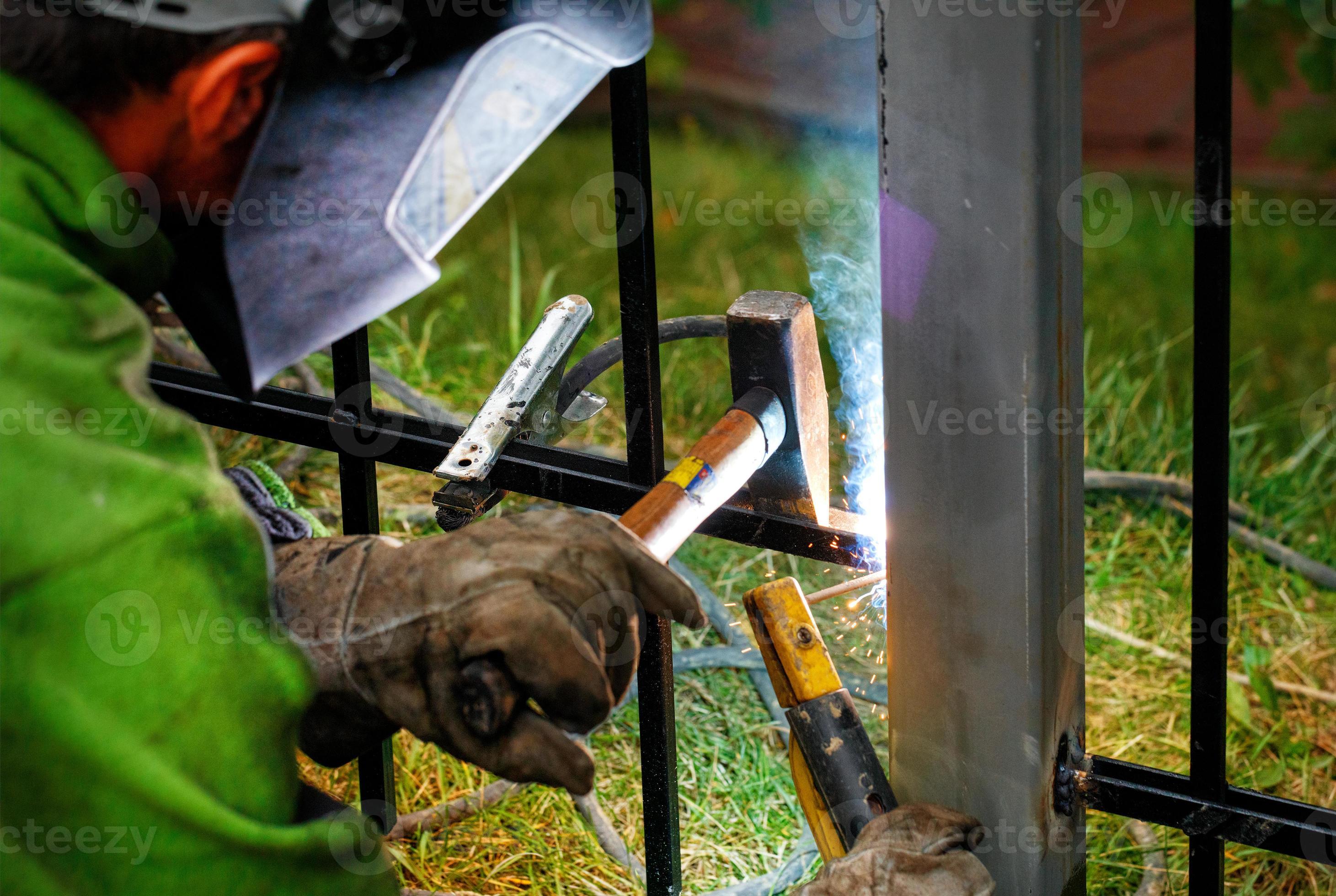 A welder wearing a protective mask welds an iron fence to a metal post