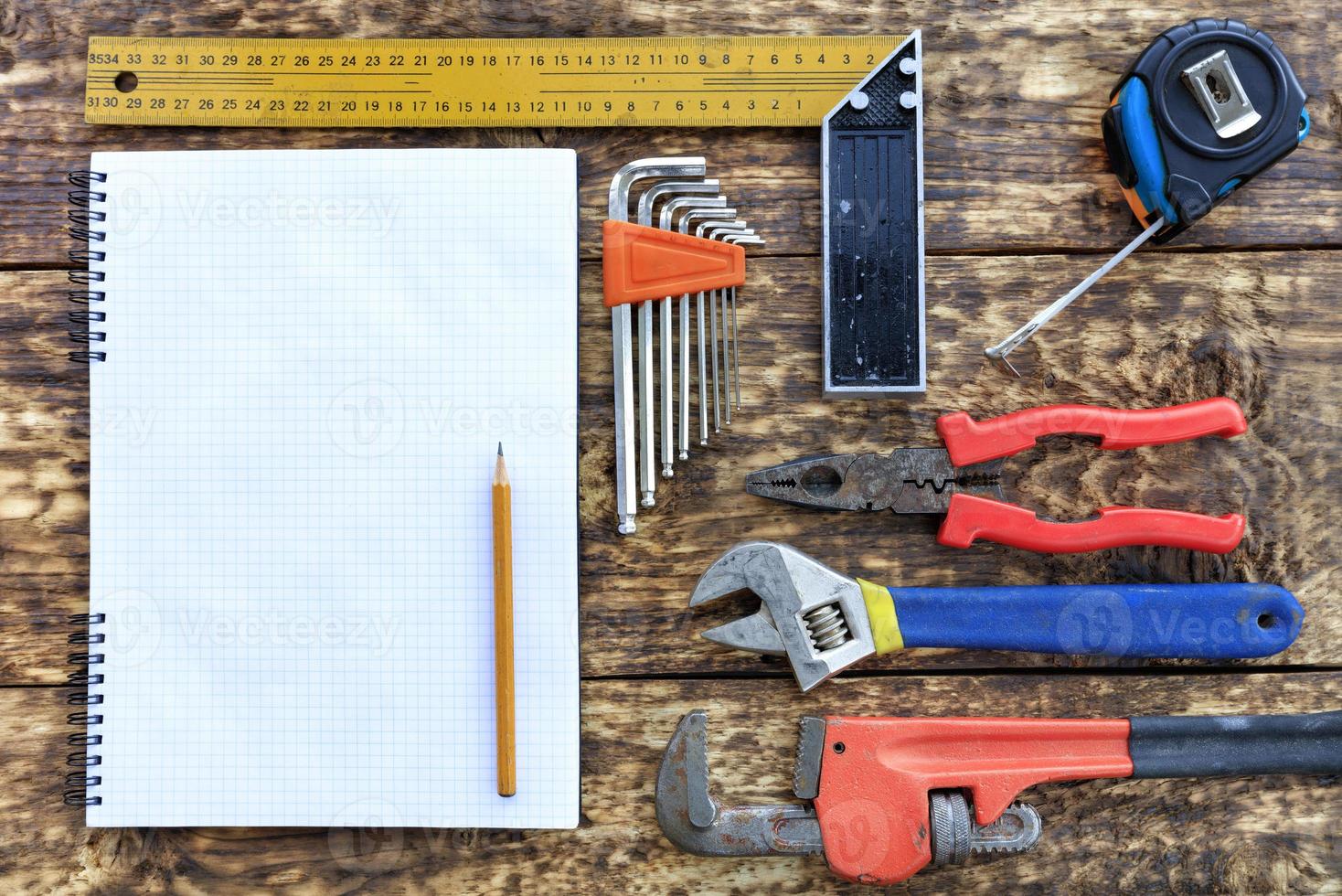 Various hand tools and blank notepad with pencil on old wooden background. photo