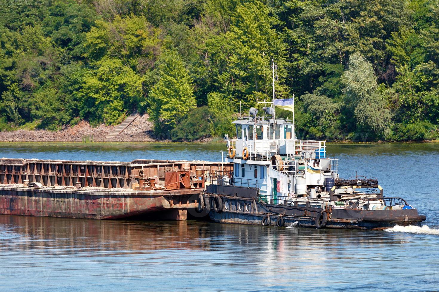 An old river tug pushes an empty barge ahead of itself along the river bank. photo