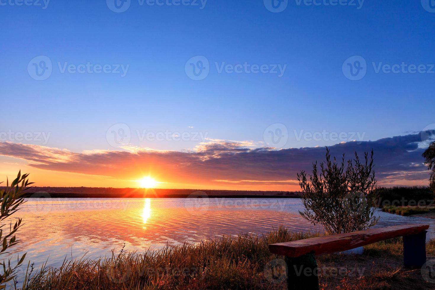 The bright rising sun rises above the horizon behind a wide river and is reflected in a wavy water surface. photo