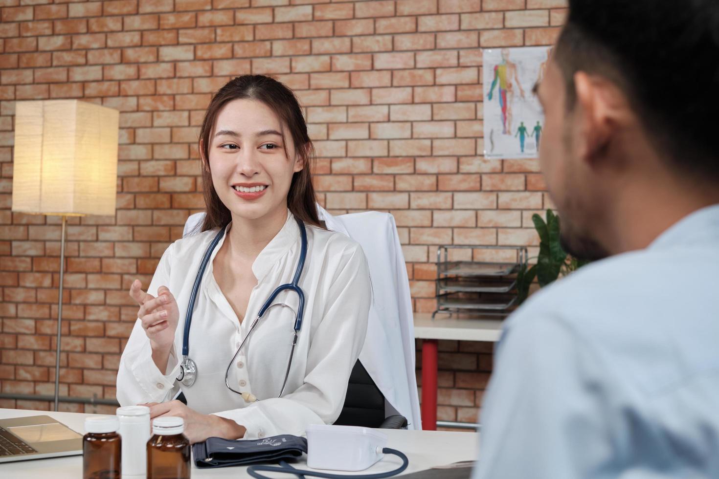 Beautiful woman doctor in white shirt who is Asian person with stethoscope is health examining male patient in brick wall background medical clinic, smiling advising medical specialist occupation. photo