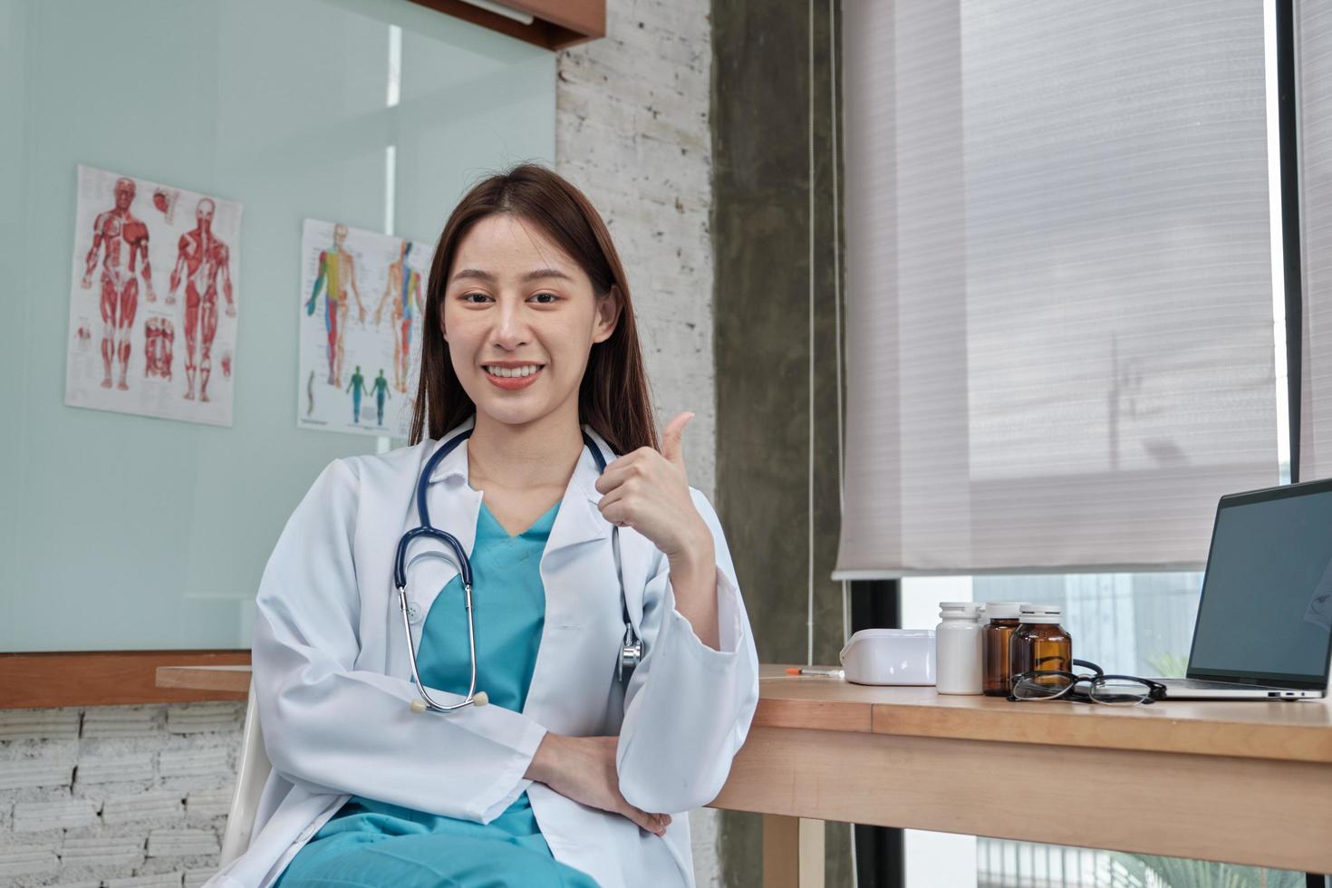 Retrato de hermosa doctora de origen asiático en uniforme con estetoscopio, pulgar hacia arriba, sonriendo y mirando a la cámara en una clínica, una persona con experiencia en tratamiento profesional. foto