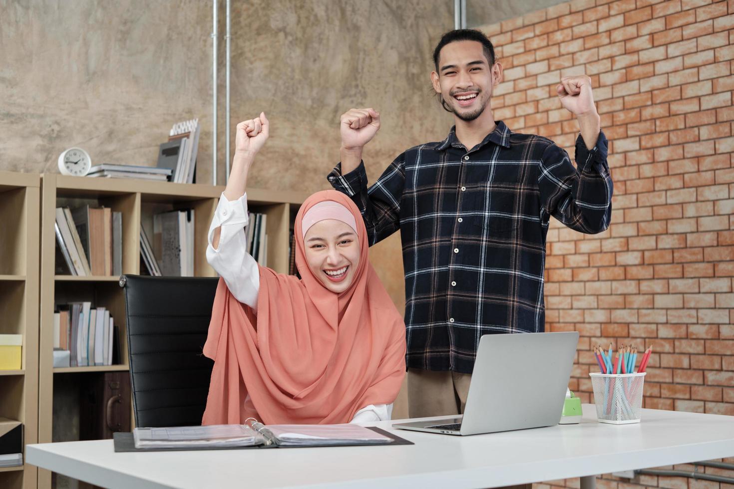 Young startup partners who are Islamic people stand, raise their hands to congratulate the success of small office e-commerce businesses. Two couple colleagues smile and are happy working. photo