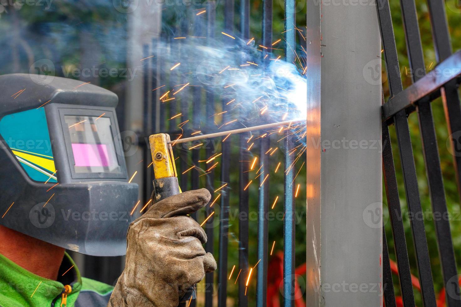 A welder in a protective helmet and gloves works with metal, welds a metal fence in a park area. photo