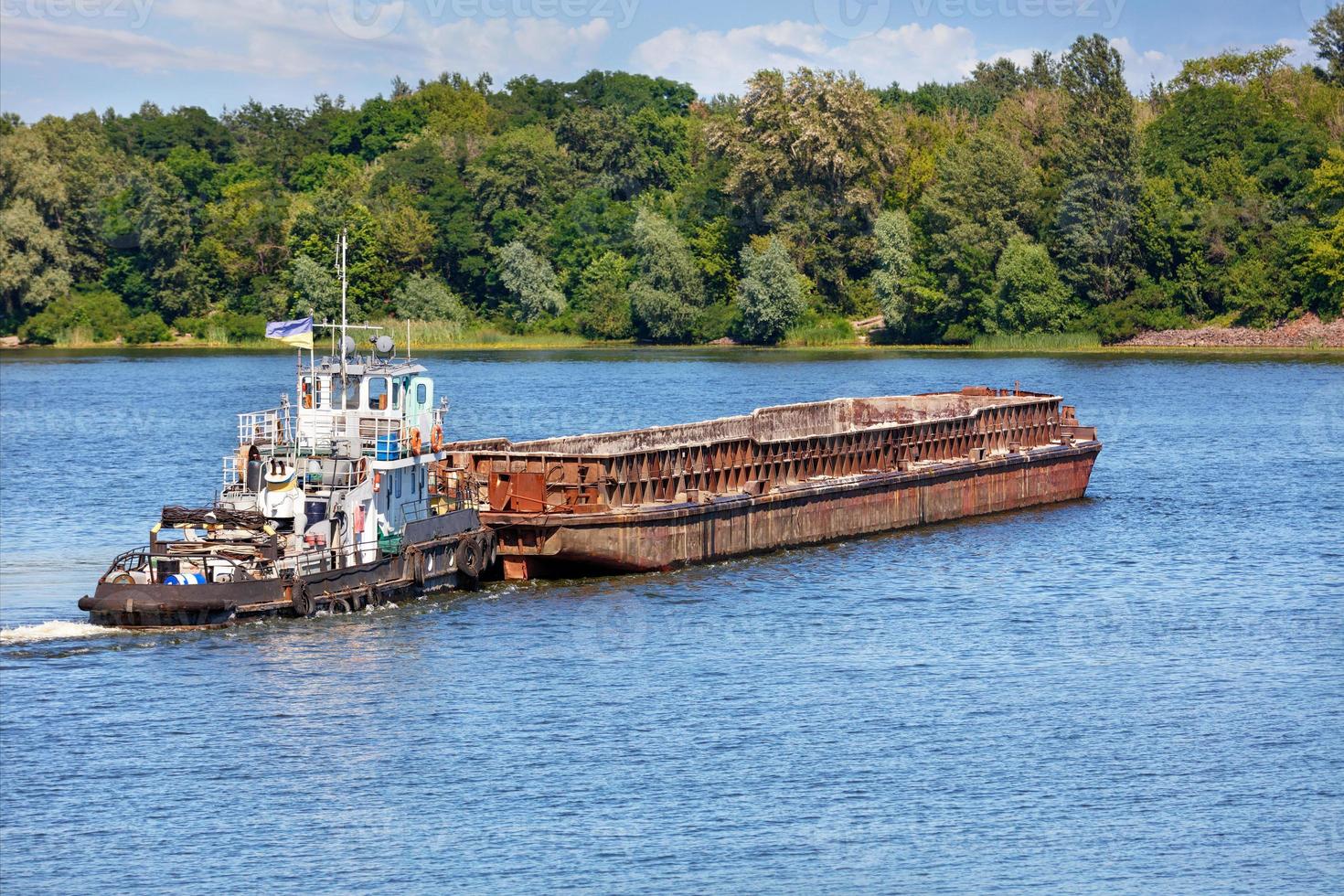 A river tugboat with an empty rusty barge goes crosses a wide river. photo