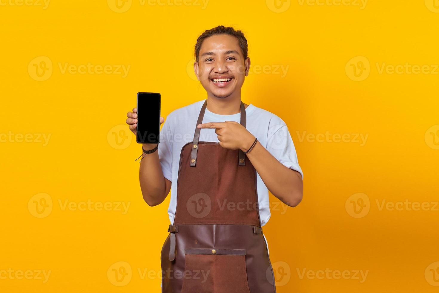 Sonriente joven guapo apuntando a la pantalla del teléfono inteligente en blanco sobre fondo amarillo foto