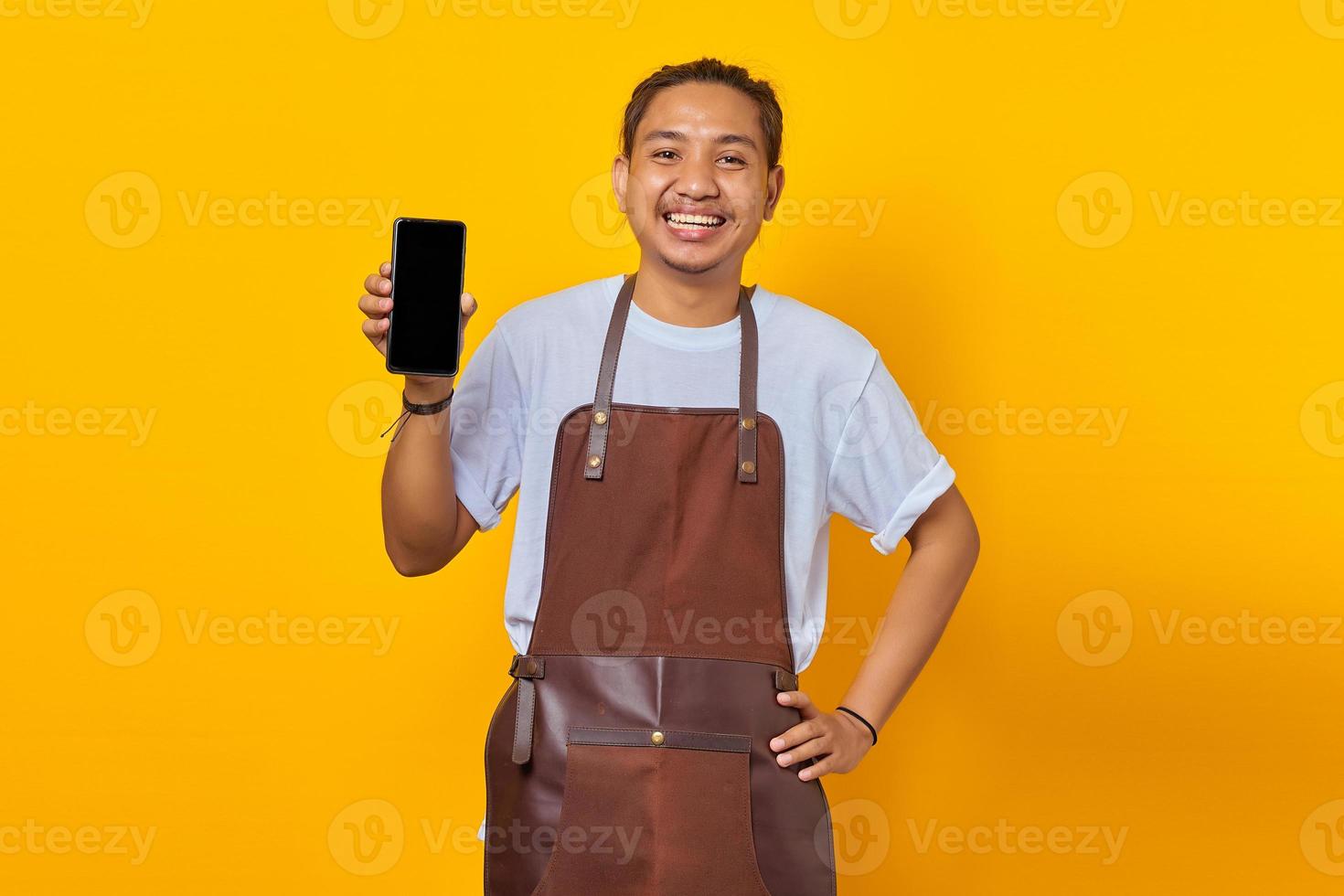 Cheerful asian barista showing smartphone blank screen over yellow background photo