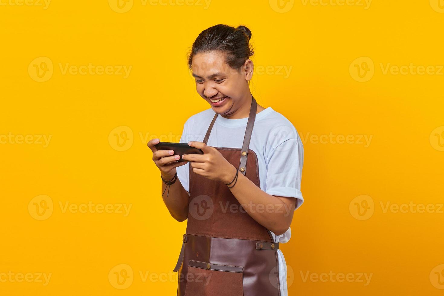 Portrait of happy young Asian barista playing video game on mobile isolated on yellow background photo