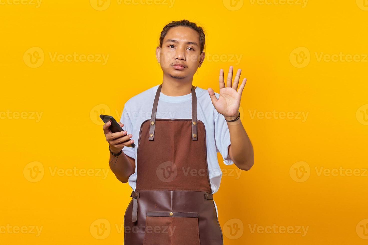 Portrait of Asian young man wearing surprised apron and doing stop gesture with palms on yellow background photo