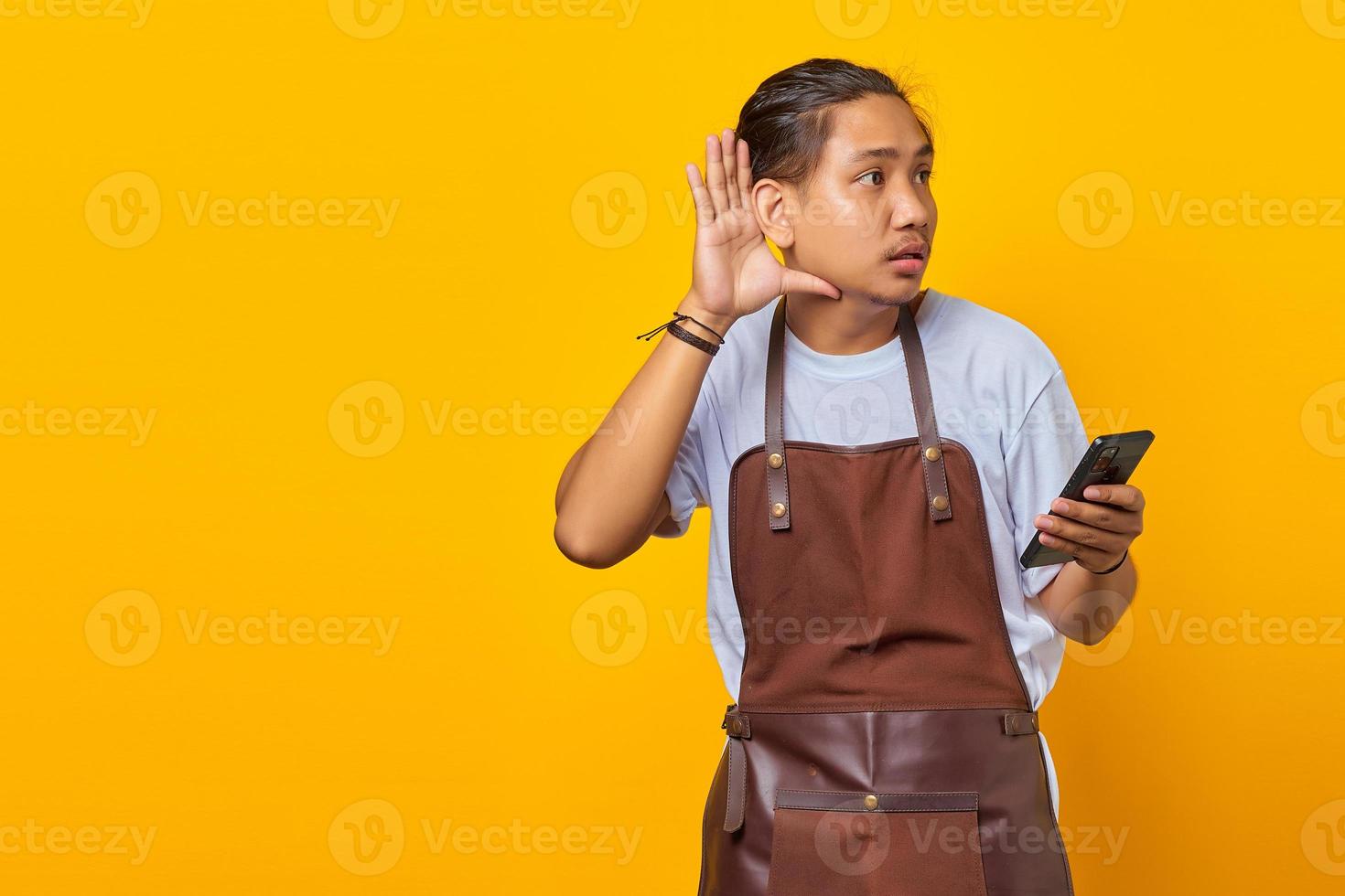 Portrait of serious looking Asian man trying to overhear secret conversation and holding smartphone photo