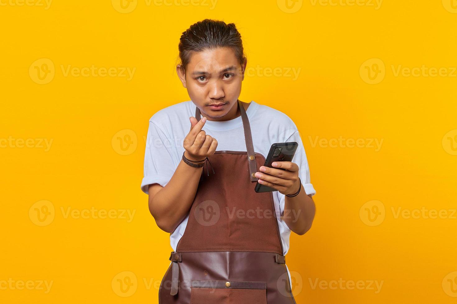 Portrait of cheerful Asian young man showing finger heart and holding smartphone photo