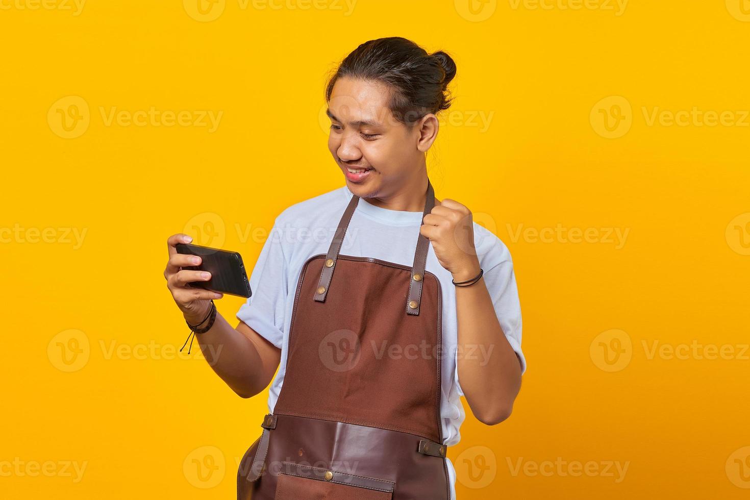 Portrait of cheerful young Asian man playing video game on mobile and celebrating success on yellow background photo