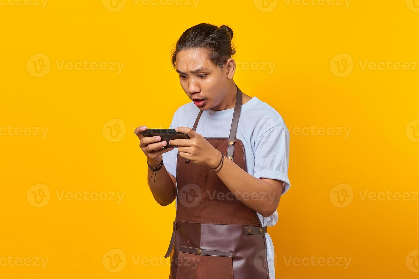 Portrait of handsome asian man wearing apron looking annoyed while playing game on smartphone photo