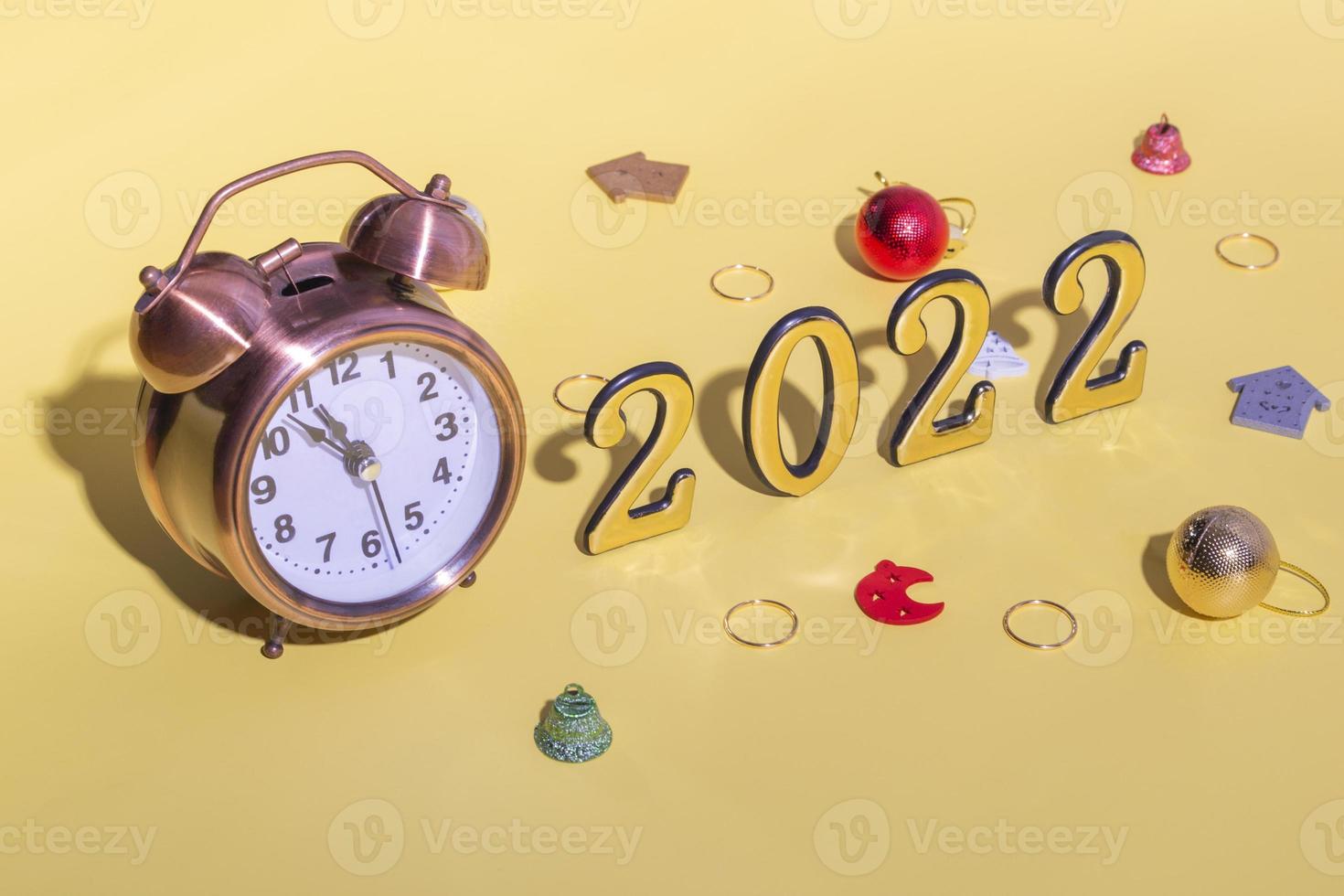 Alarm clock, Christmas decor and numbers 2022 with hard shadow on yellow background. New year celebration minimal concept. Close up, copy space photo