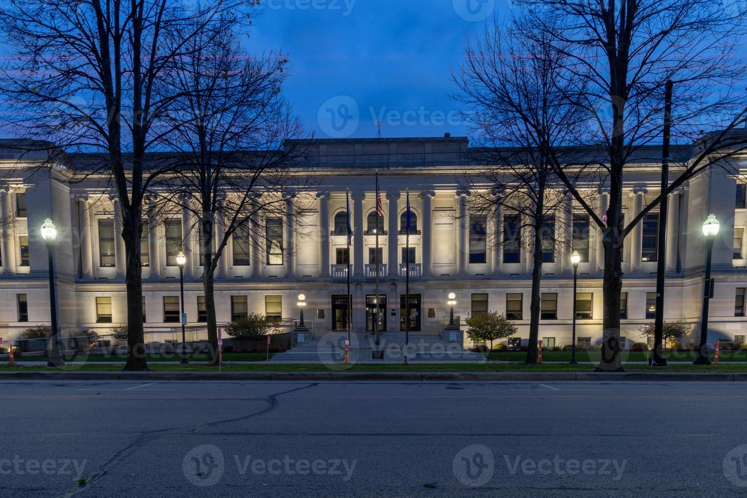 Palacio de Justicia del condado de Kenosha, Wisconsin, iluminado por la noche. altos y majestuosos árboles desnudos que bordean la entrada. Grandes escalones que conducen a la entrada. foto