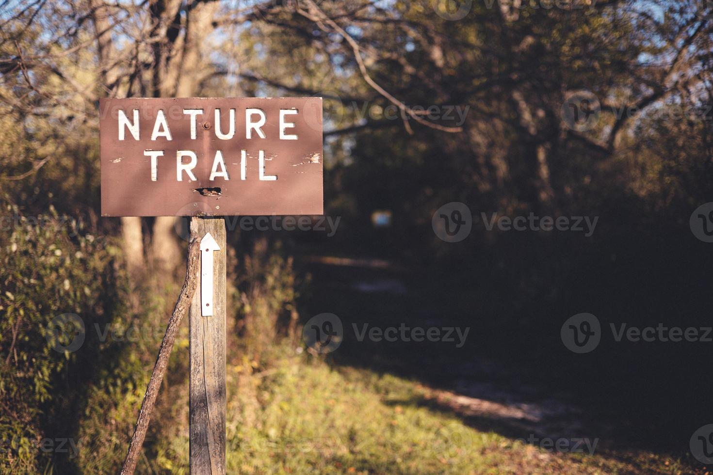 Wooden sign marking trail name on a wooden post. Arrow marking which direction the trail goes. Sun shining through the tree branches casting shadows on the ground. photo