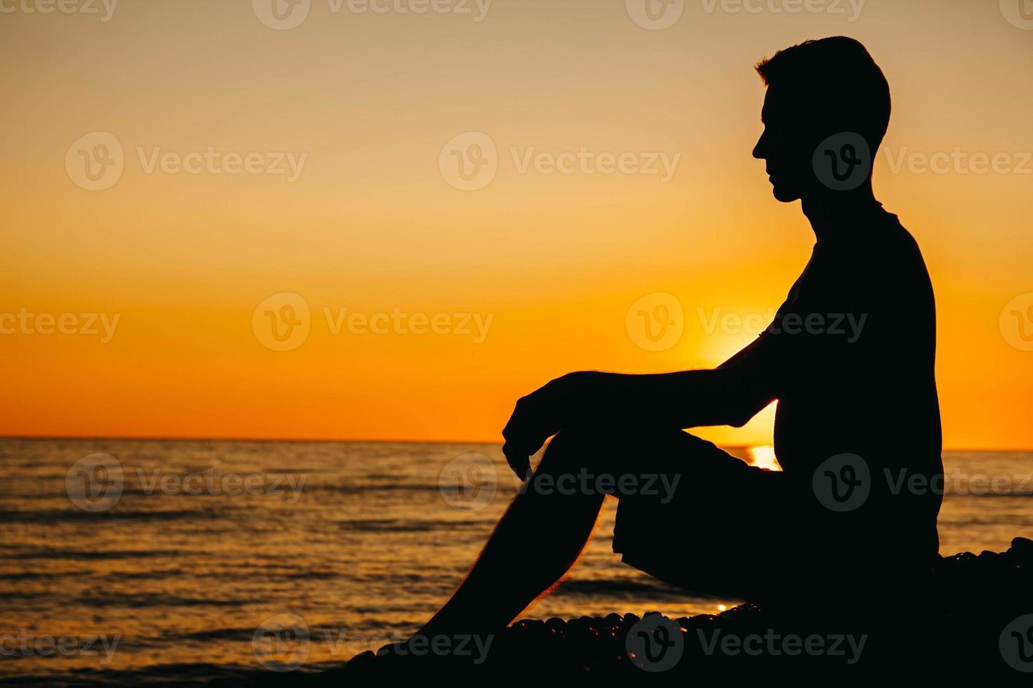 Silhouette of a man sitting and contemplating on beach on sea background and sunset photo