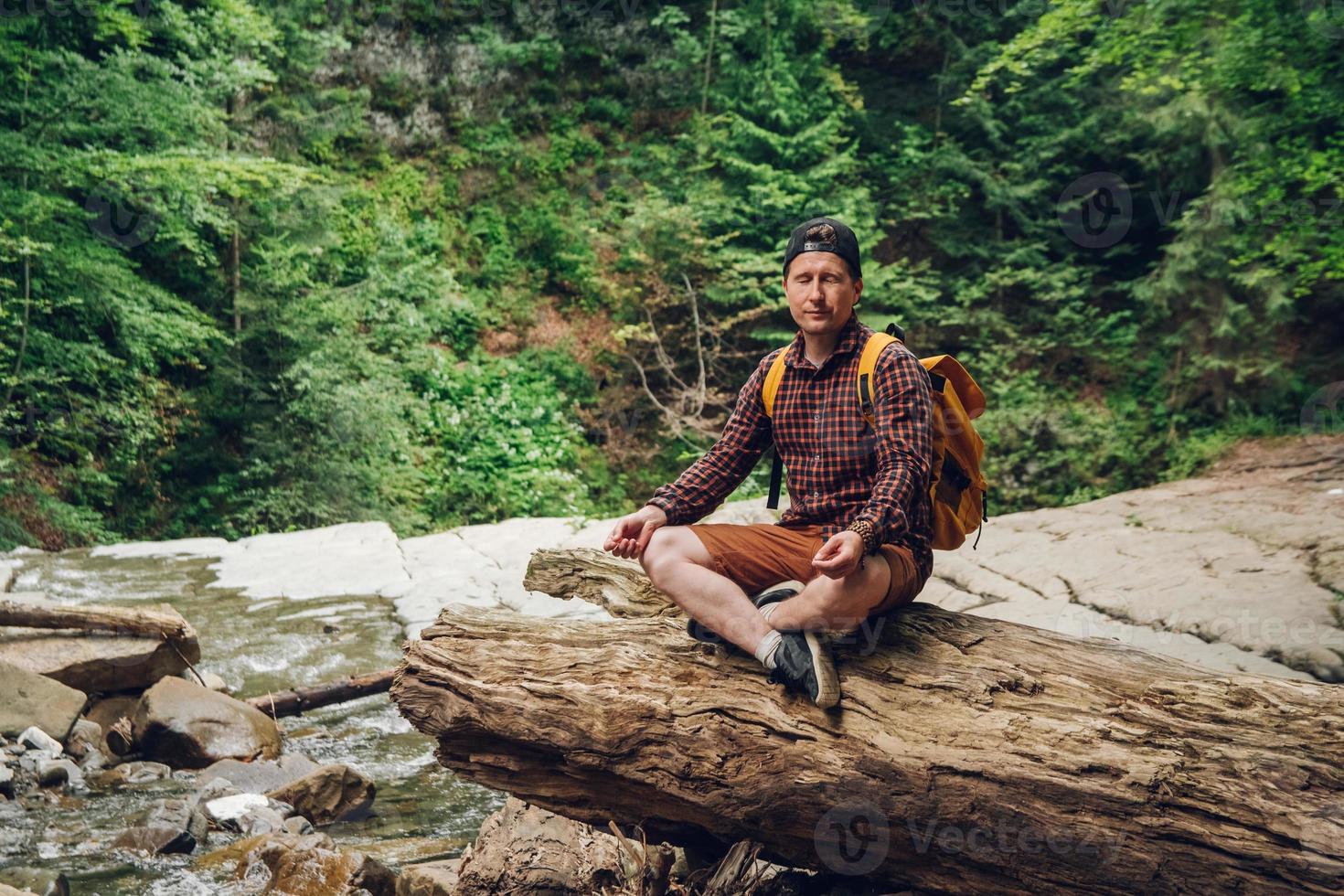 Man in a meditative position with a backpack sitting on a tree trunk against background of forest and river photo