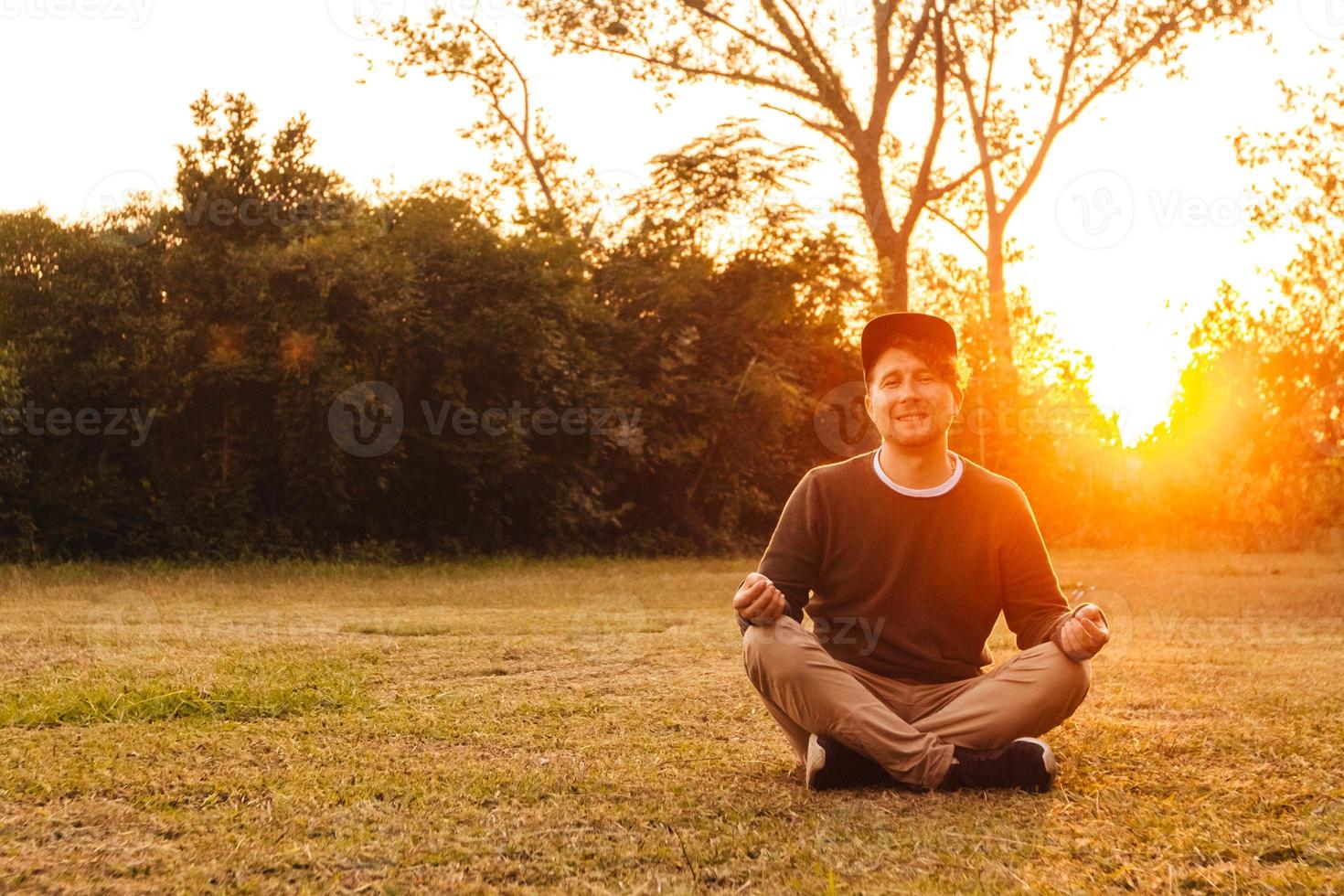 El hombre en una posición meditativa se sienta en un césped sobre un fondo de bosque y puesta de sol foto