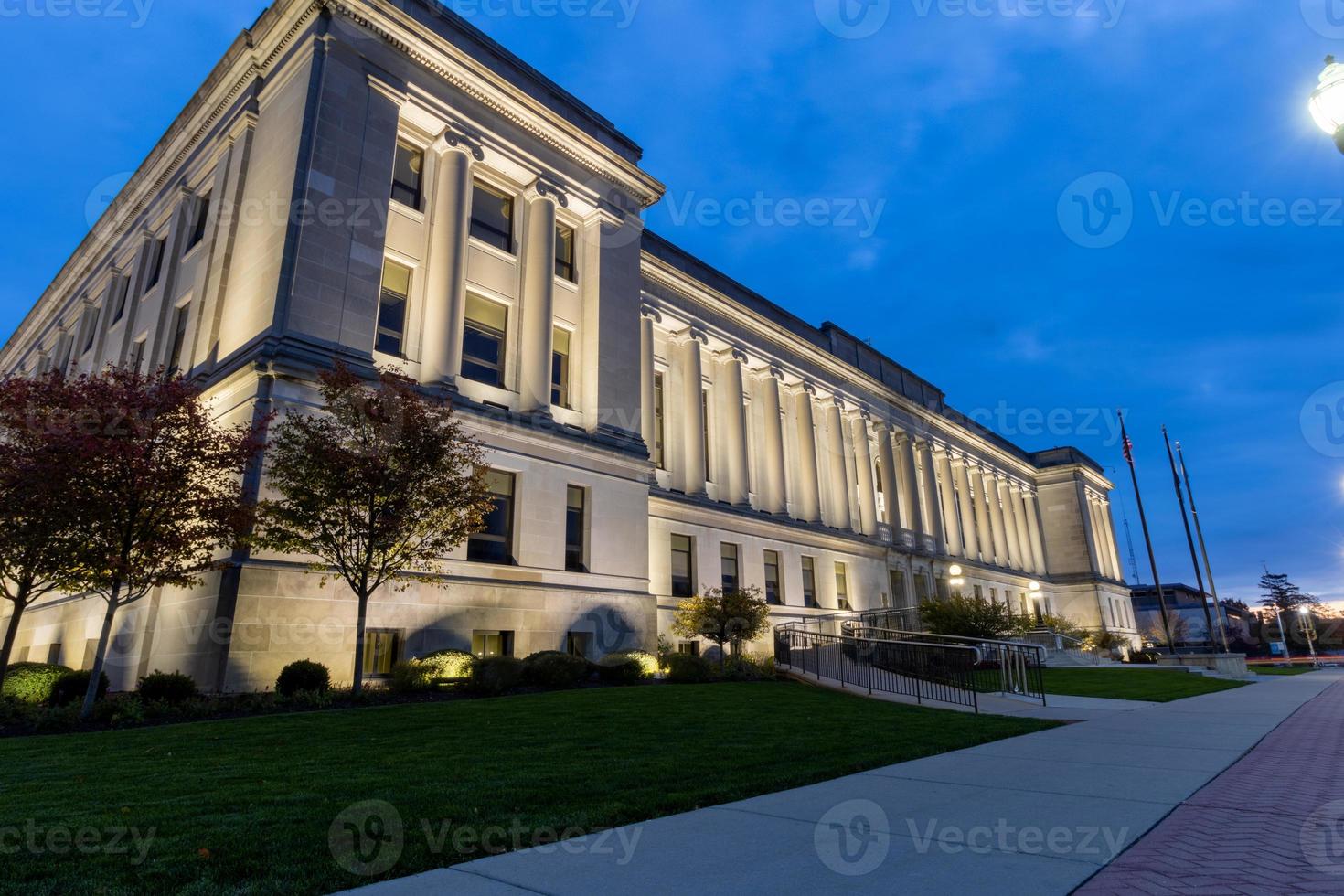 Kenosha, Wisconsin, County Courthouse at night. Brilliant blue sky.  Courthouse lights shining on the architecture.  Wide sidewalk lined with bricks and street lights. Tree branches glowing in light photo