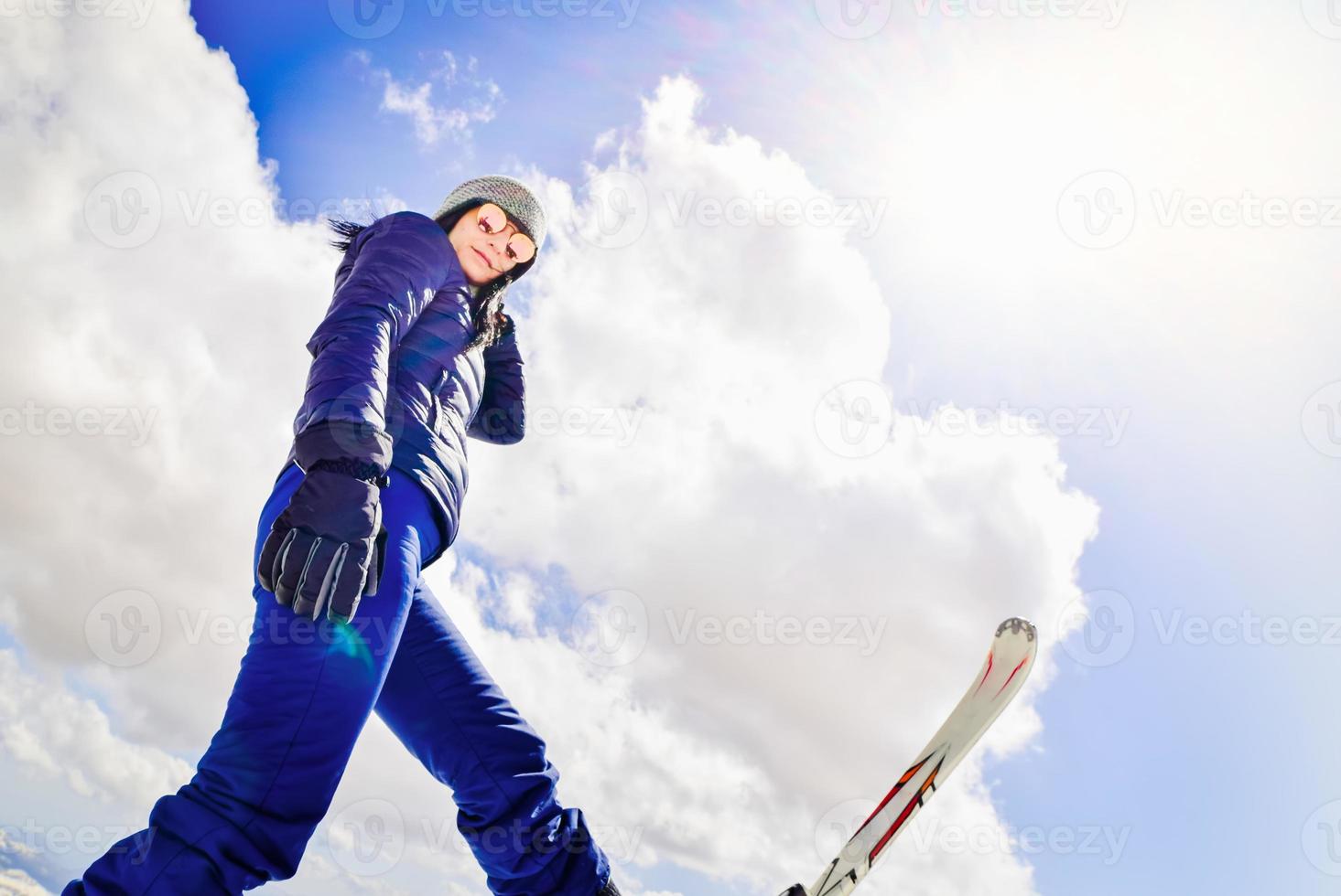 Happy excited cool caucasian brunette woman with skies pose in winter outfit in mountains. Copy paste vertical background top left photo