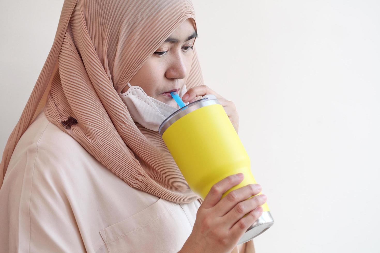 Chica musulmana abre una mascarilla quirúrgica para beber agua sobre fondo pastel. concepto de coronavirus covid-19. foto
