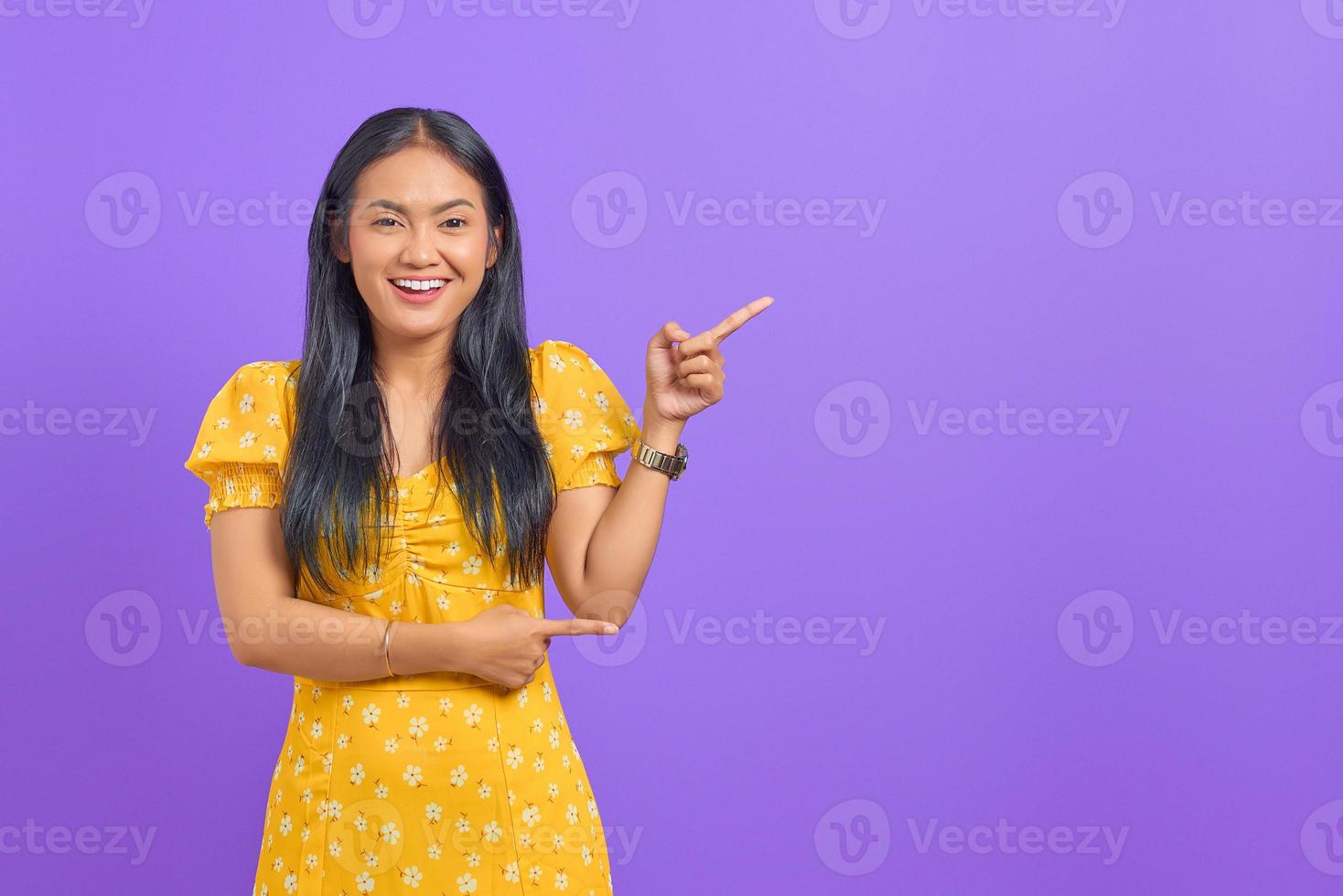 Portrait of smiling young Asian woman pointing finger at copy space on purple background photo