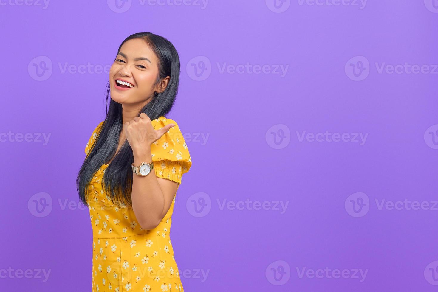 retrato, de, sonriente, joven, mujer asiática, señalar con el pulgar, en, copia, espacio, en, fondo púrpura foto