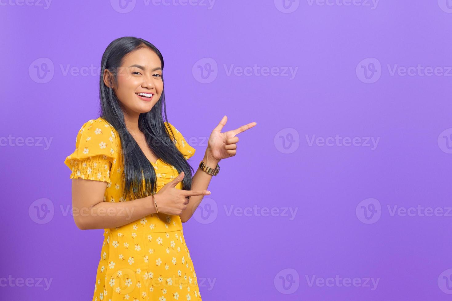 Portrait of smiling young Asian woman pointing finger at copy space on purple background photo